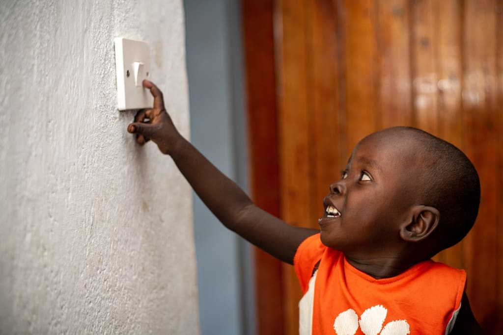 3-year-old Thokozani reaching on to the light bulb switch. He reaches his arm up to a white textured wall and puts his finger on the switch as he smiles looking at it. He is wearing an orange sleeveless shirt, and a wooden panel door is behind him.