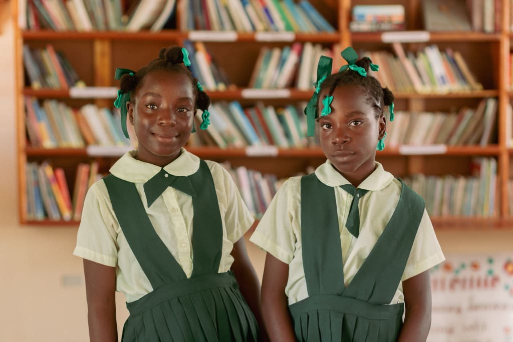 Portrait of Carolie with her twin sister, Laurie. They are both wearing green and white uniforms and have green barrettes in their hair. There are bookshelves filled with books in the background.