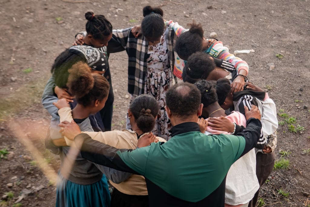 A group of youth standing in a circle. They are embracing each other and looking to the ground.