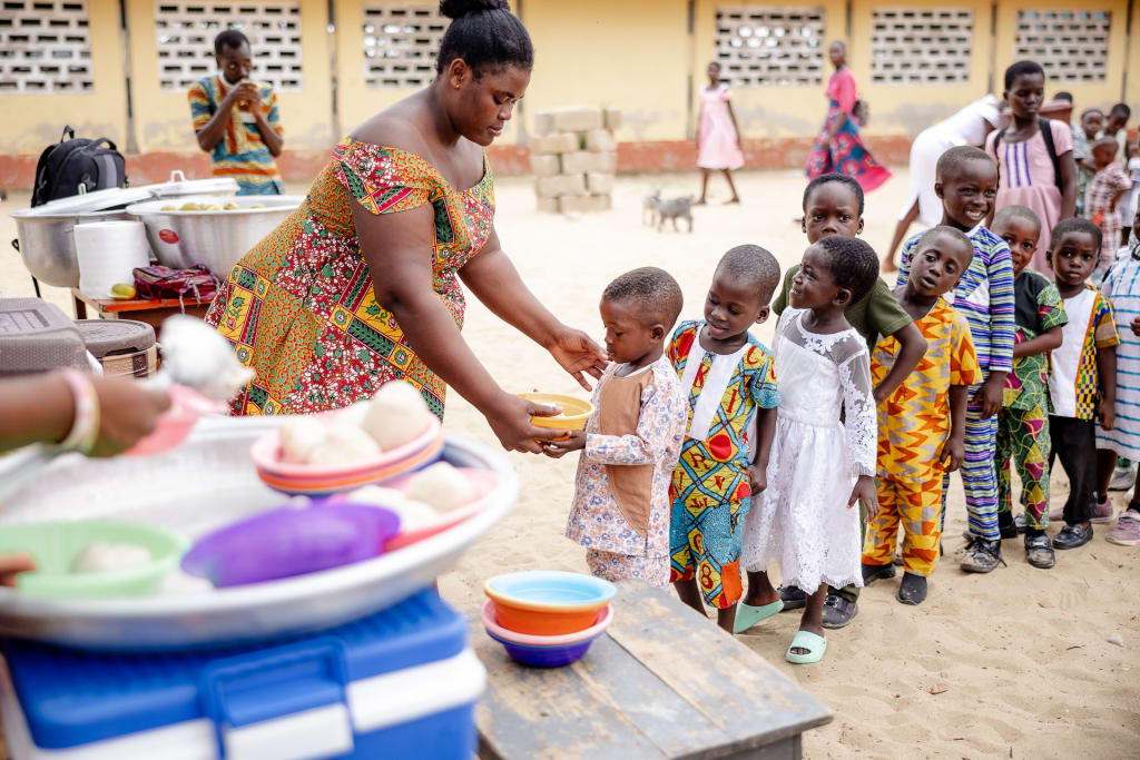 A tutor hands a young participant a bowl of breakfast. Several others wait patiently in line. Breakfast is served under giant palm trees on sand.