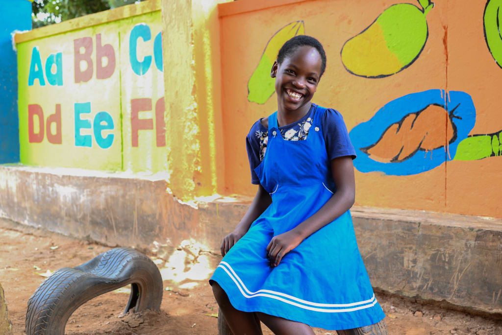 Juliet is smiling as she sits outside on a playground posing for a picture. There is a bright wall behind her with part of the alphabet painted on it.