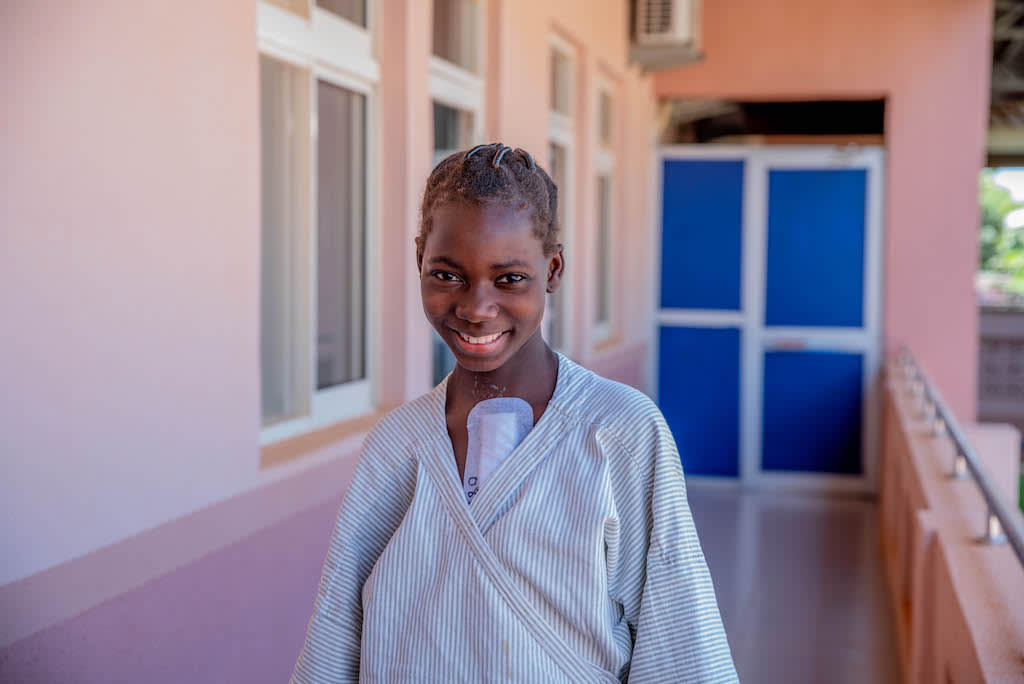 Françoise is standing outside the National Hospital in a hospital gown with a white bandage on her chest.