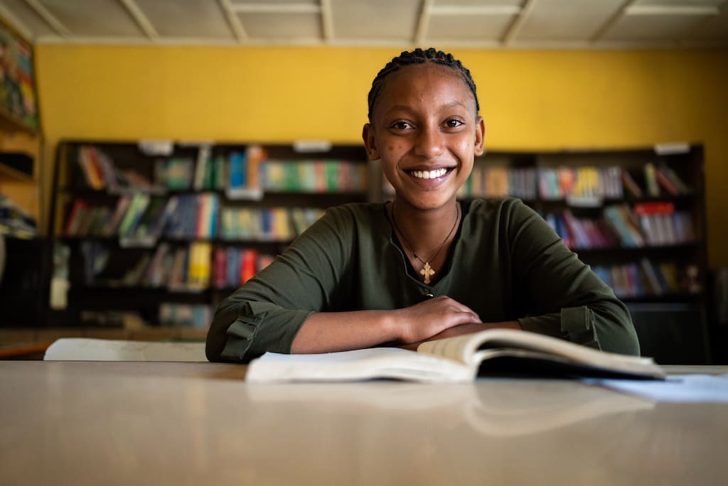 Mart is an advocate against child marriage. She is wearing a green dress. She is sitting at a table inside the centre. There is a book in front of her. Behind her are shelves full of books.