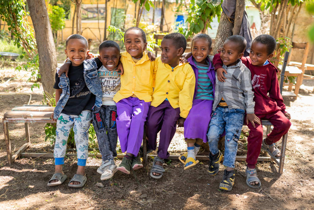 A group of seven children sit on a bench with their arms around each other and smile.
