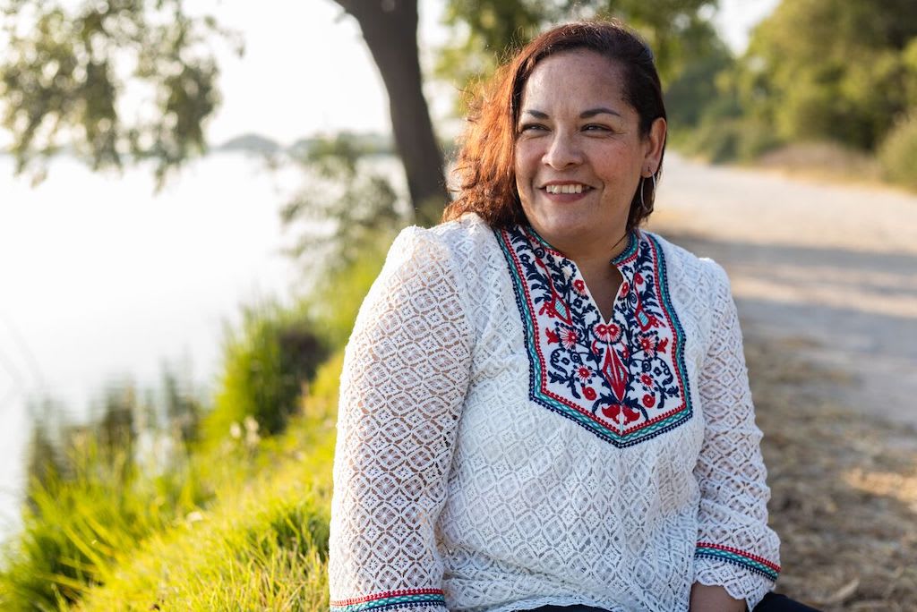 A Mexican woman wearing a white blouse sits by a body of water.