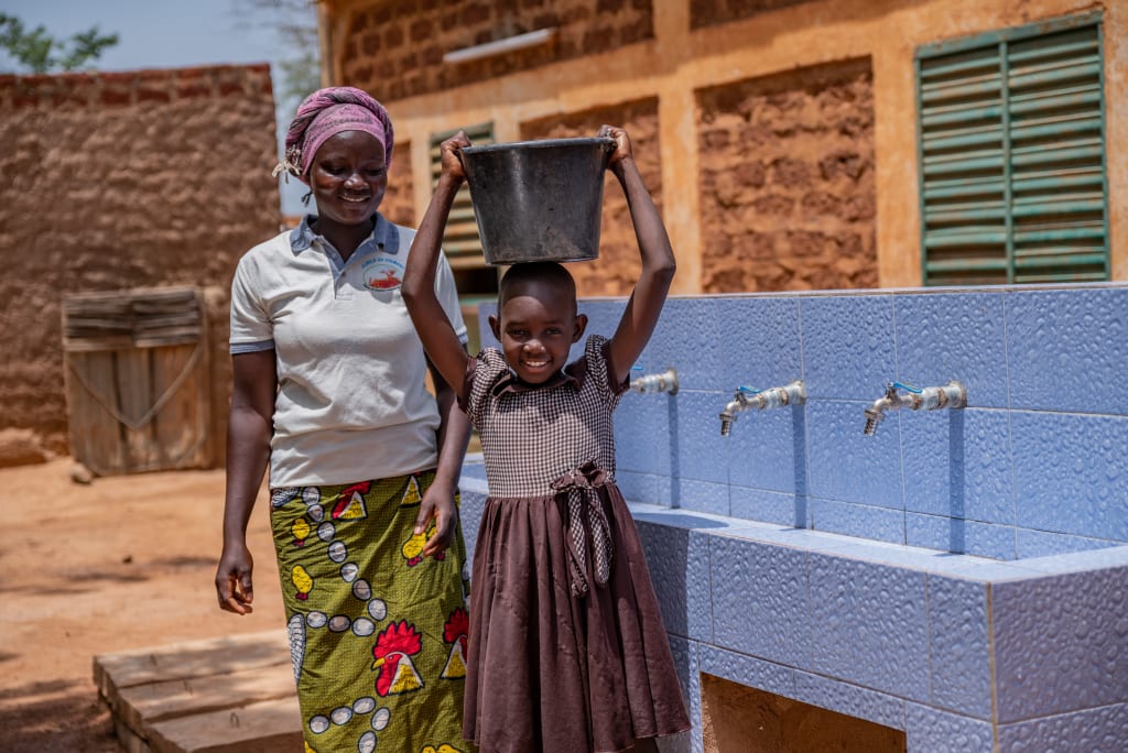 A girl in a brown dress stands smiling with a bucket on her head in front of a block of blue sinks. A lady in a white shirt and green skirt stands smiling behind her.