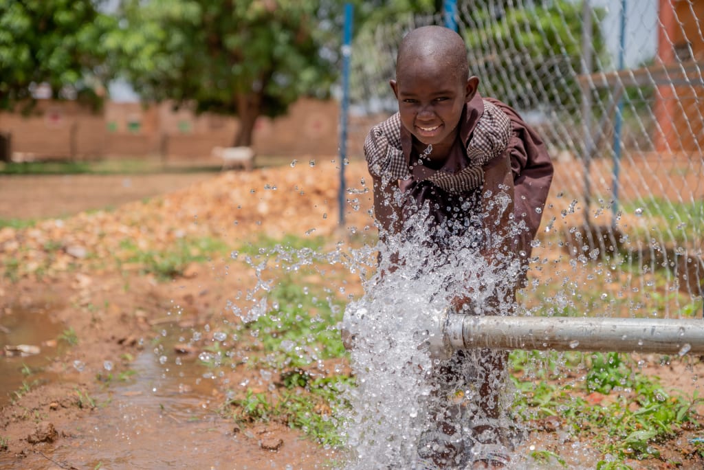 A girl in a brown dress stands in front of a pipe that is bursting with water.
