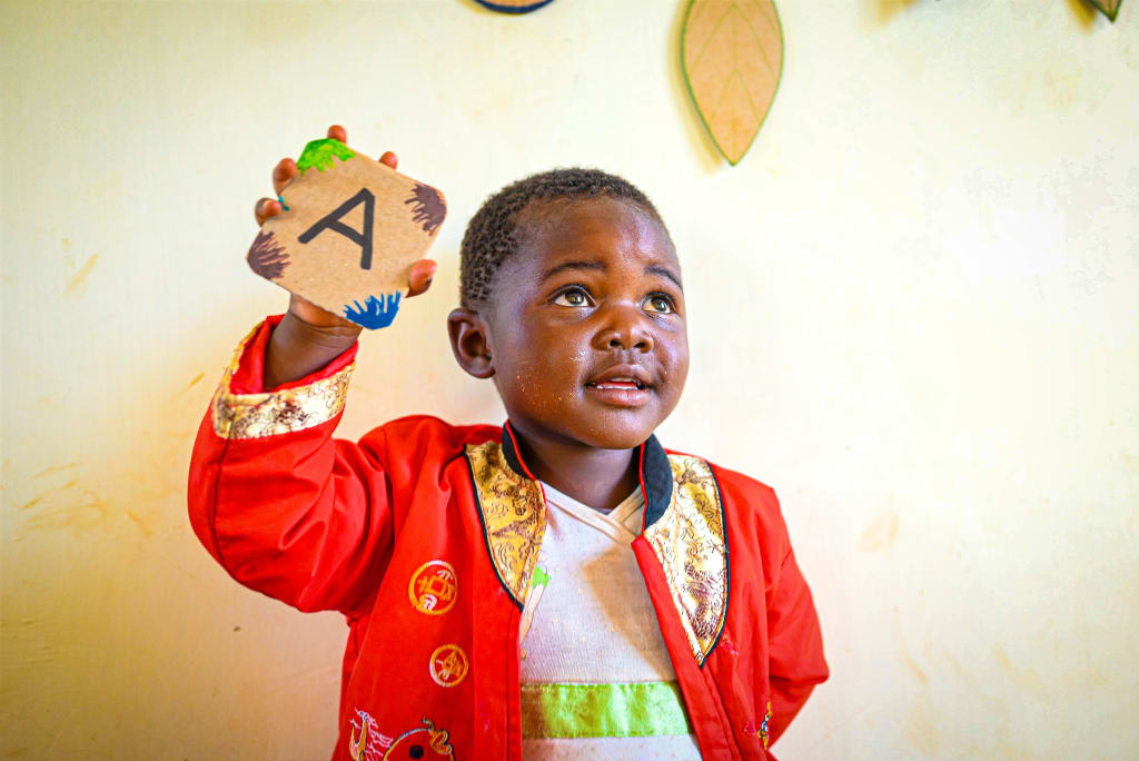 A little girl in a red coat holds up a tile with the letter A on it.