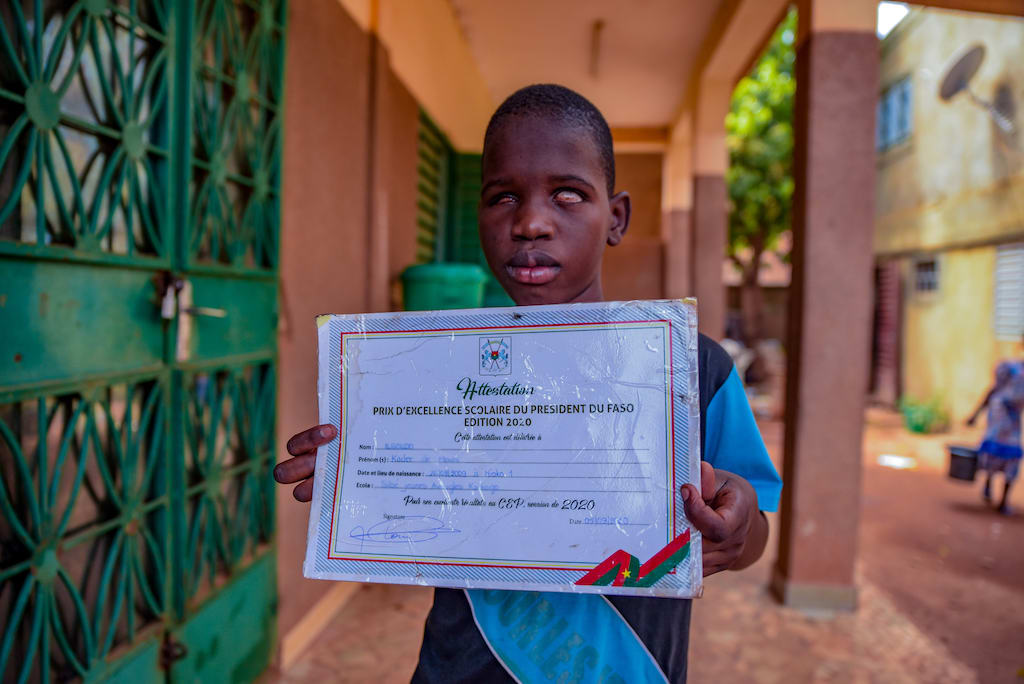 Kader is wearing a blue shirt. He is standing outside his school and is holding up a certificate he received from the president. Kader's school is brown and has green doors.
