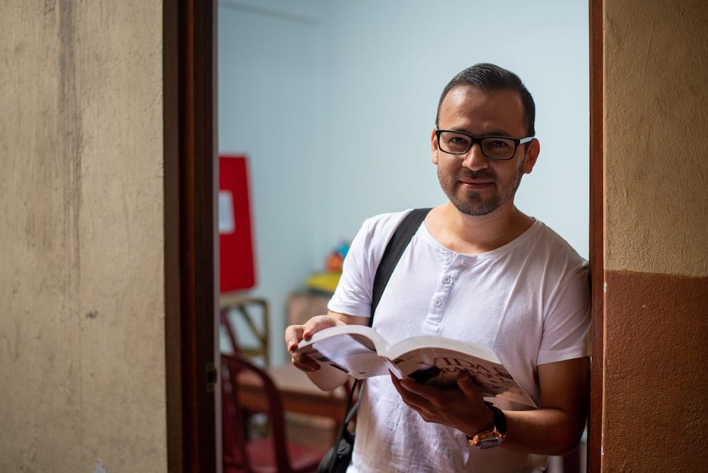 John leans in a doorway, with an open book in his hands.
