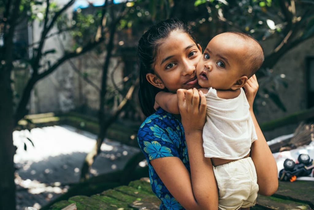 A girl wearing a blue flowered dress hold a baby close to her. There are trees behind them.