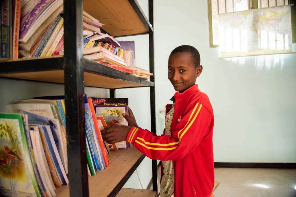 Abel, in a red jacket, is standing next to a bookshelf touching some of the books.