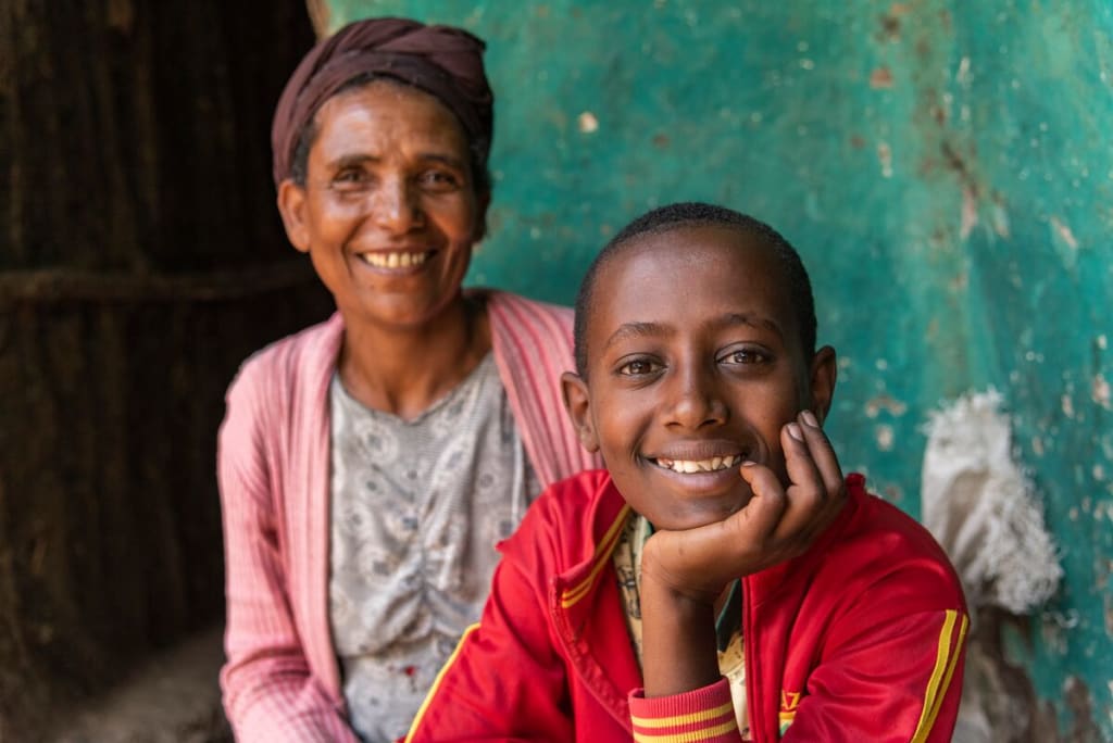 Abel, in a red jacket, is at home sitting and posing for a picture with his mom, Astede, who is wearing a pink sweater. The wall behind them is green.