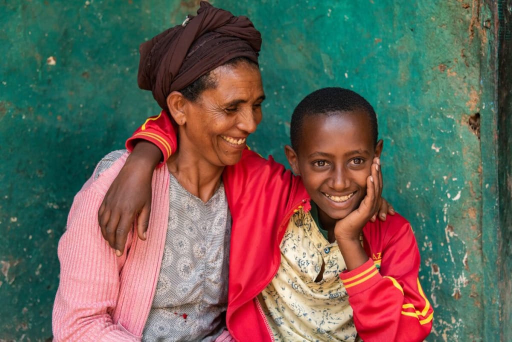 Abel, in a red jacket, is at home sitting and posing for a picture with his mom, Astede, who is wearing a pink sweater. She is looking at him and smiling, and he has his arm around her shoulders. The wall behind them is green.