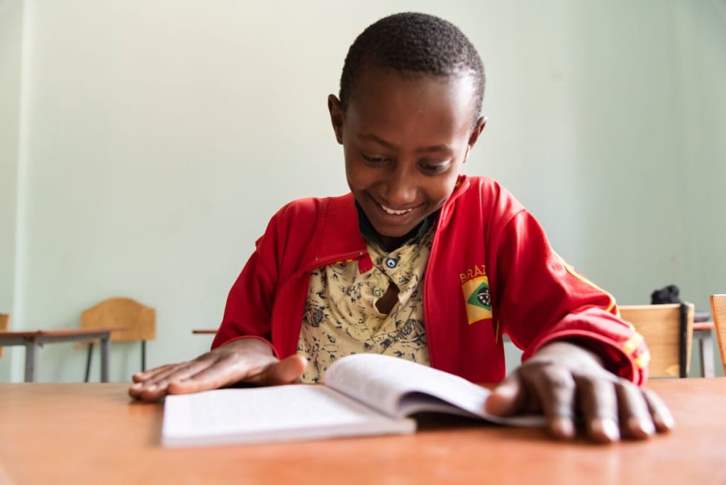 Abel, in a red jacket, is sitting at a table smiling and reading a book.