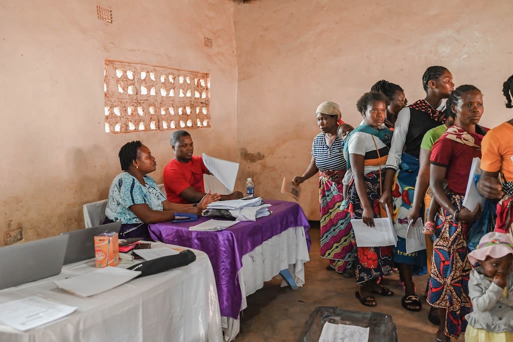 Parents are registering their children in the program. There are two white tables with women holding papers, and parents lining up.