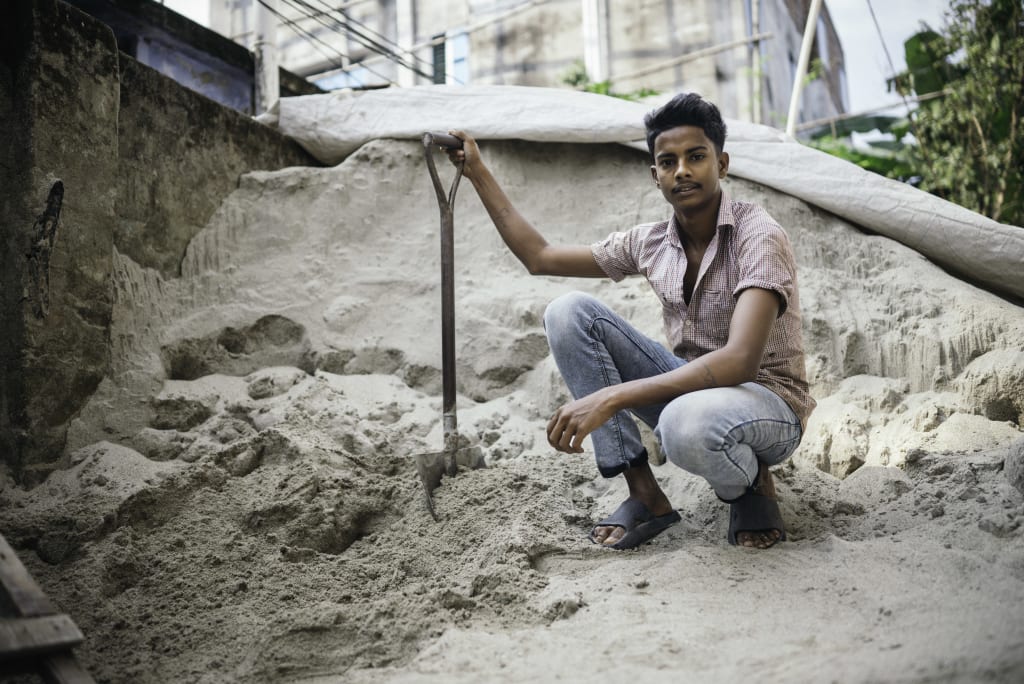 Shanto holds a shovel and kneels in a pile of sand.