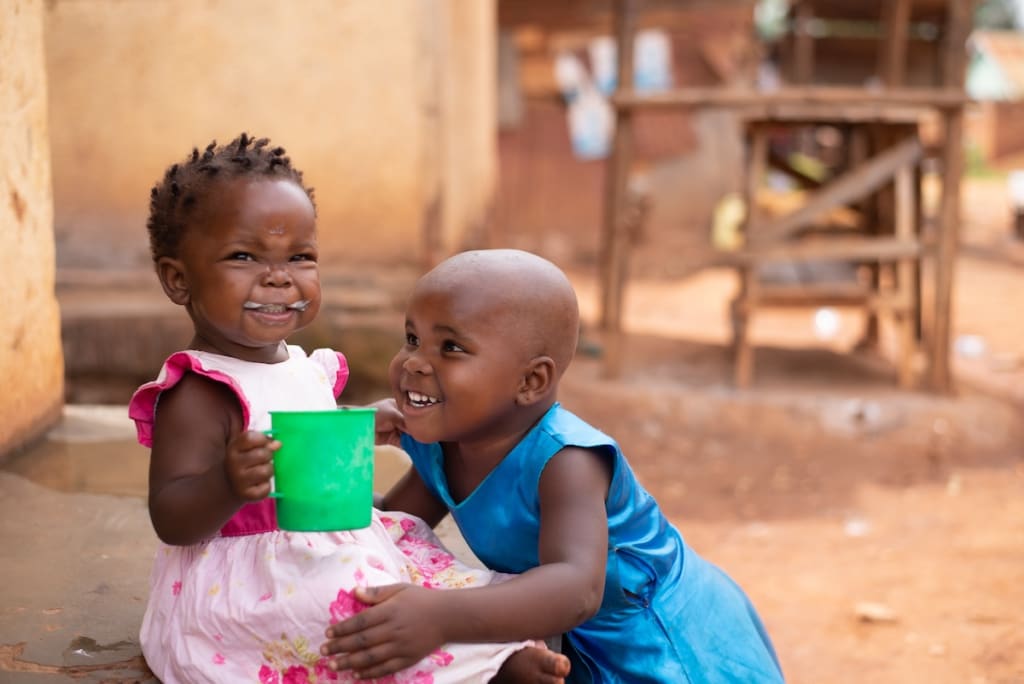 In Uganda, Charity, wearing blue, plays with her sister Shanita, wearing pink. Shanita is holding a green cup with food in it.