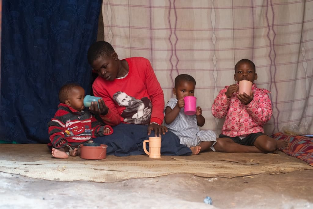 In Uganda, Aidah is wearing a red shirt and blue skirt. She is sitting down outside her home with her three children. They are eating and drinking out of cups.