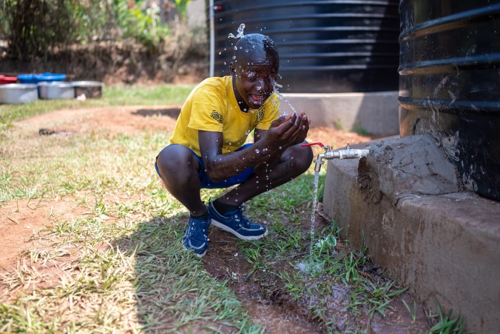 Patrick, a 13-year-old boy, squats in front of a water tank and splashes water from the tap onto his face.