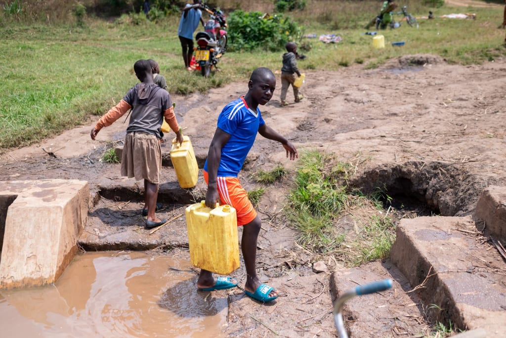 Patrick walks away from a well carrying a yellow jerrycan full of water. In the background are other children also carrying jerrycans.