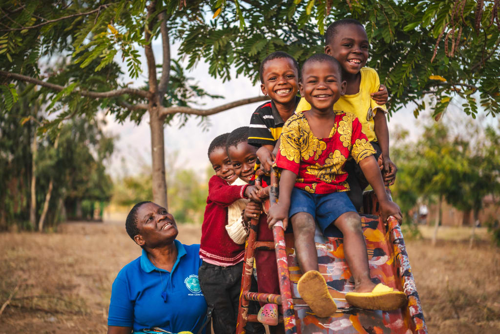 A woman in a blue shirt stands looking up at a group of children standing on the ladder of a slide.