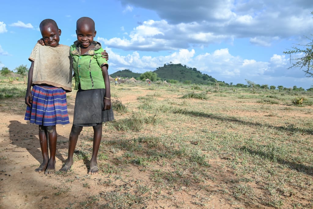 Two girls stand with their arms around each other and smile. They are standing in a big, dry field.