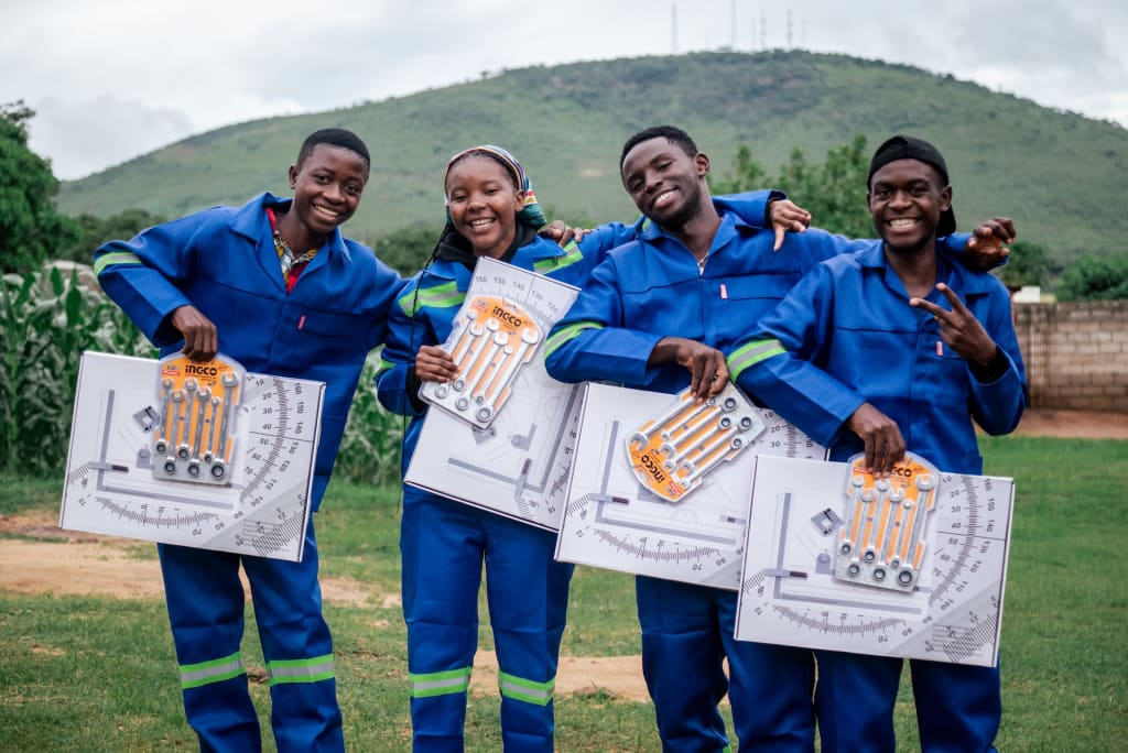 NEET beneficiaries take time to show off their equipment given to them courtesy of the front-line church and Compassion. They stand by each other with their arms around each other as they look forward to their trade school goals. They are all wearing a school uniform that consists of a blue shirt and blue pants, and they are all holding supplies for their classes. Behind them is a corn field, trees and a small mountain.