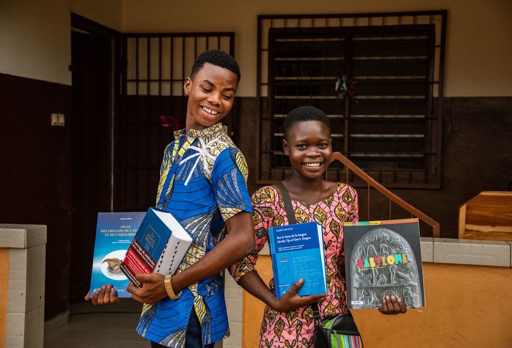 Two teens in Togo stand holding up their textbooks. They are smiling beside each other.