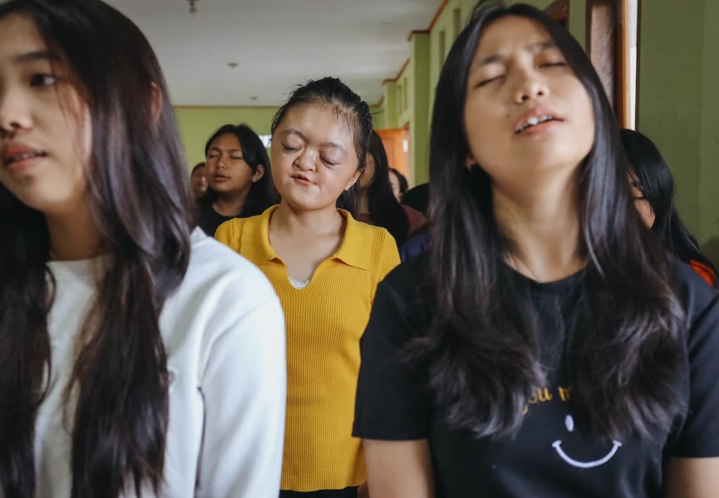 Karunia is wearing a yellow t-shirt while closing her eyes and singing. There are other girls around her singing. They're in a classroom.