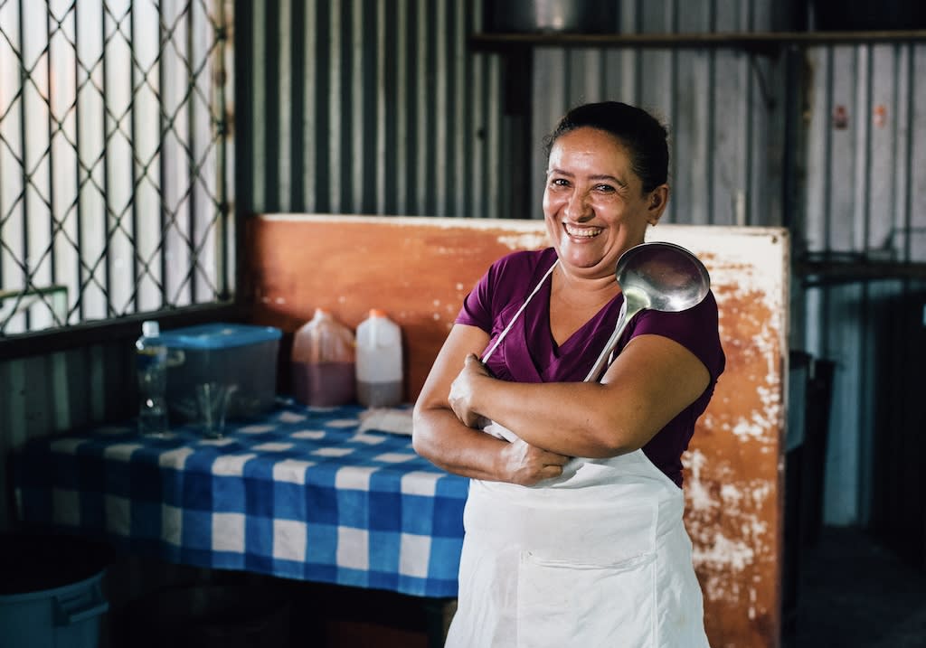 A woman smiles while crossing her arms in front of a kitchen table. She is wearing an apron and a purple shirt and is holding a ladle.