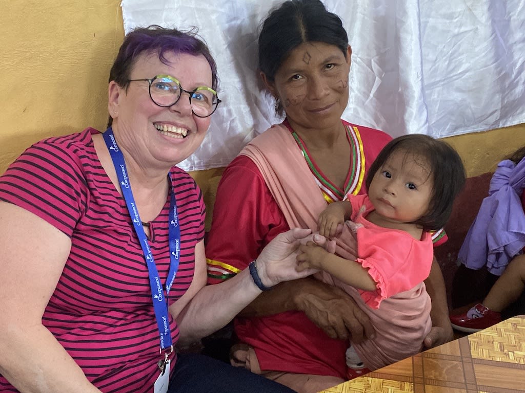 Lynn wears a pink shirt with stripes. She is holding the hand of a baby who is wearing a pink shirt. Her mother is holding her and wearing a red dress.