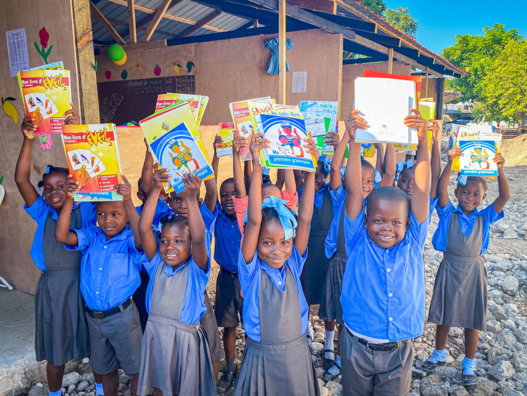 A group of children wearing blue and gray are standing outside the centre and are holding their new books up in the air.