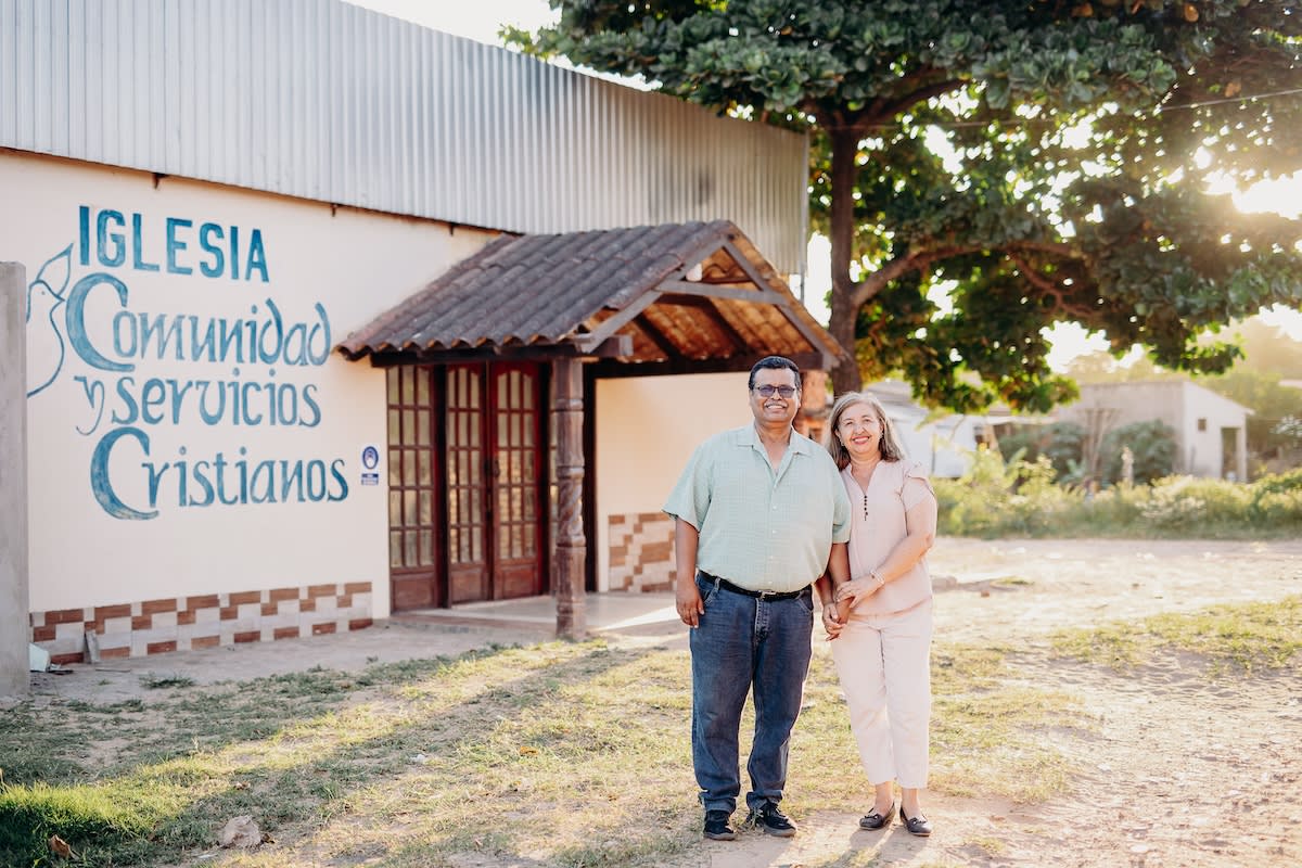 In Bolivia, Pastor Raul and Sister Graciela are standing in front of their church, Iglesia Comunidad y Servicios Cristianos. They are holding hands and smiling.