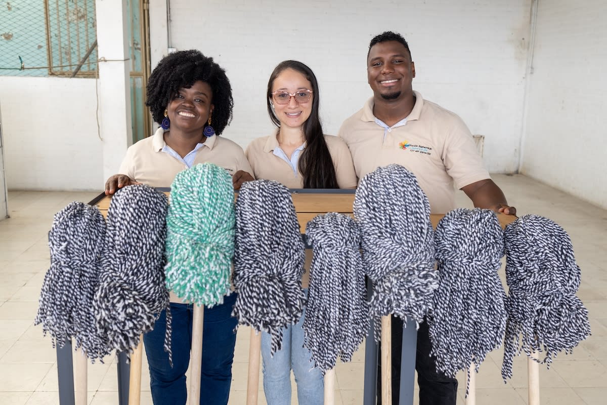 From left to right, Annie, Sandra and Carlos hold up a display of mops they have made while smiling.