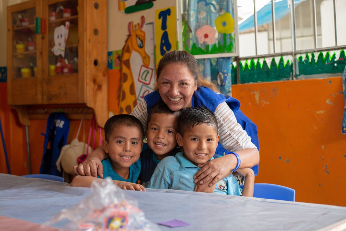 A woman in a blue vest hugs three young boys and they are all smiling