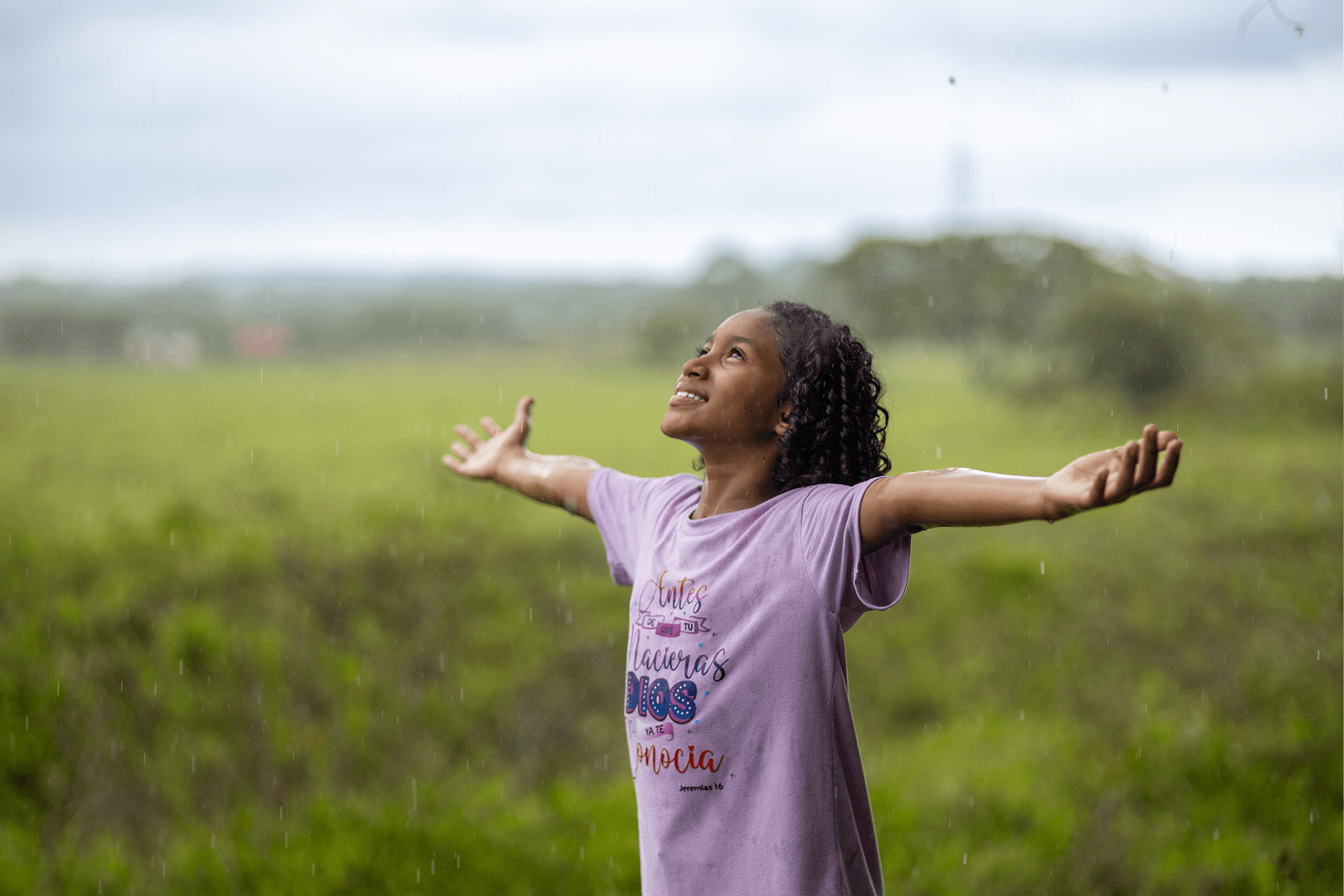 A little girl in a purple t-shirt stands in a field with her hands outstretched and looks to the sky.