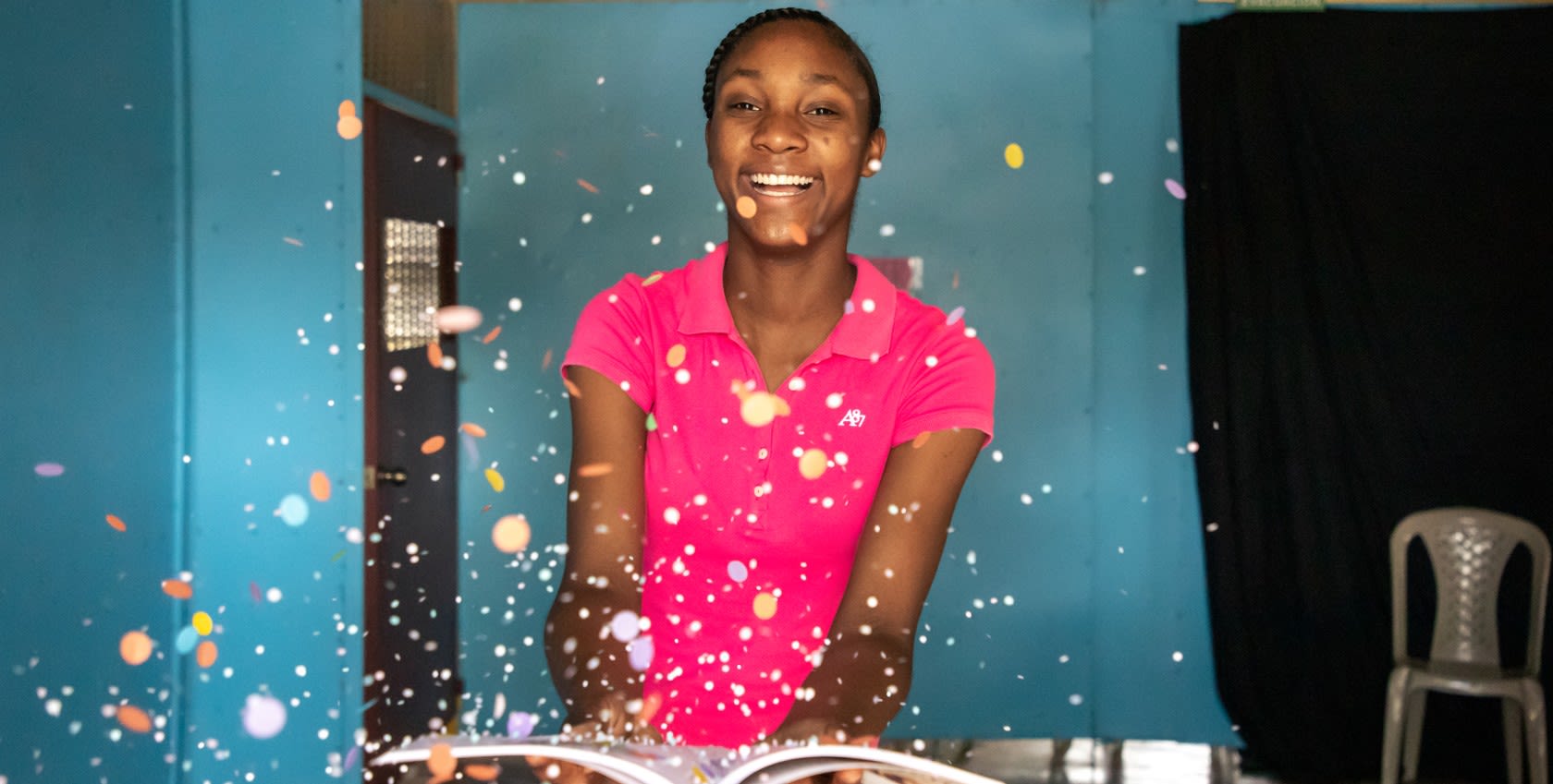 A girl wearing a pink shirt, holding an open book and smiling happily.