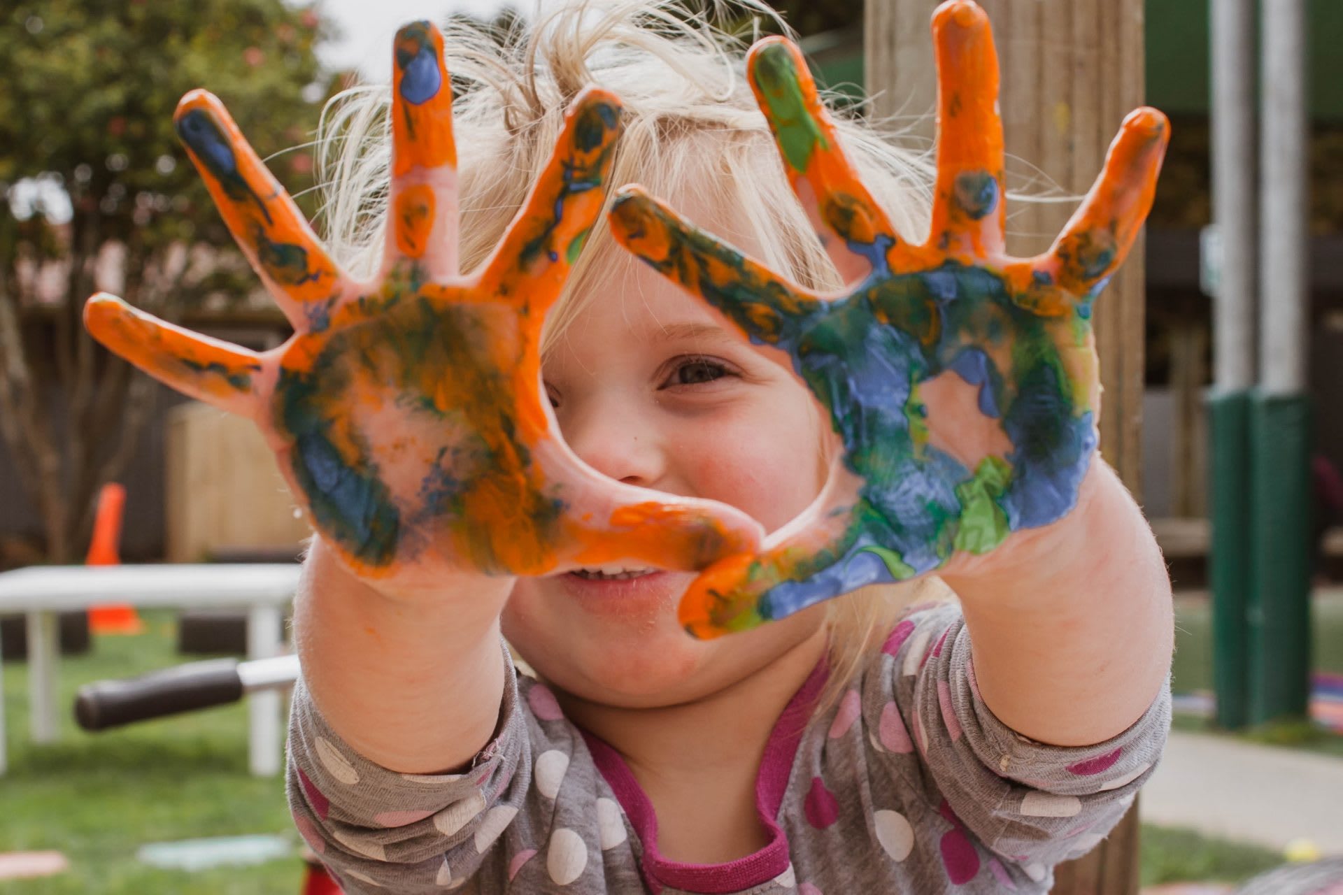 Little girl holds up her hands that are covered with blue and orange paint. She is smiling.