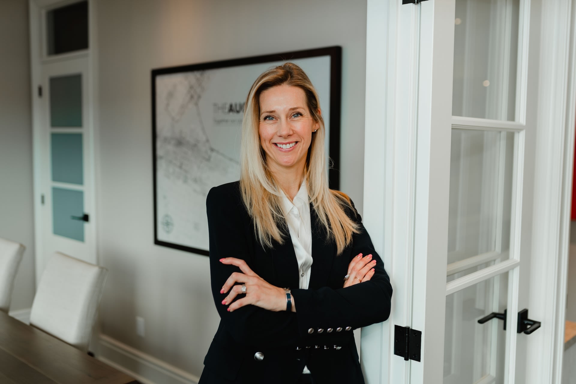 A portrait of Aimee in an office boardroom. She is standing, wearing a black blazer and has her arms crossed.