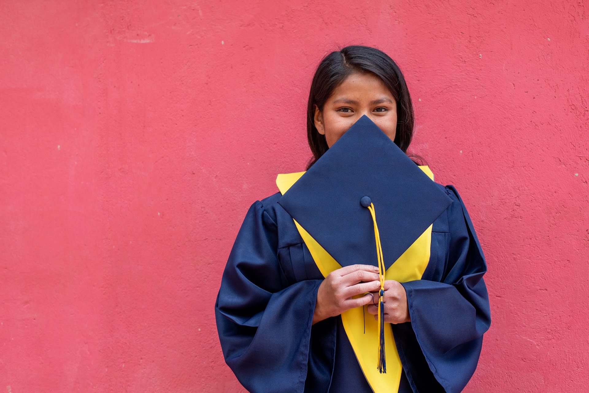A girl in a graduation gown holds her graduation cap up to her face.