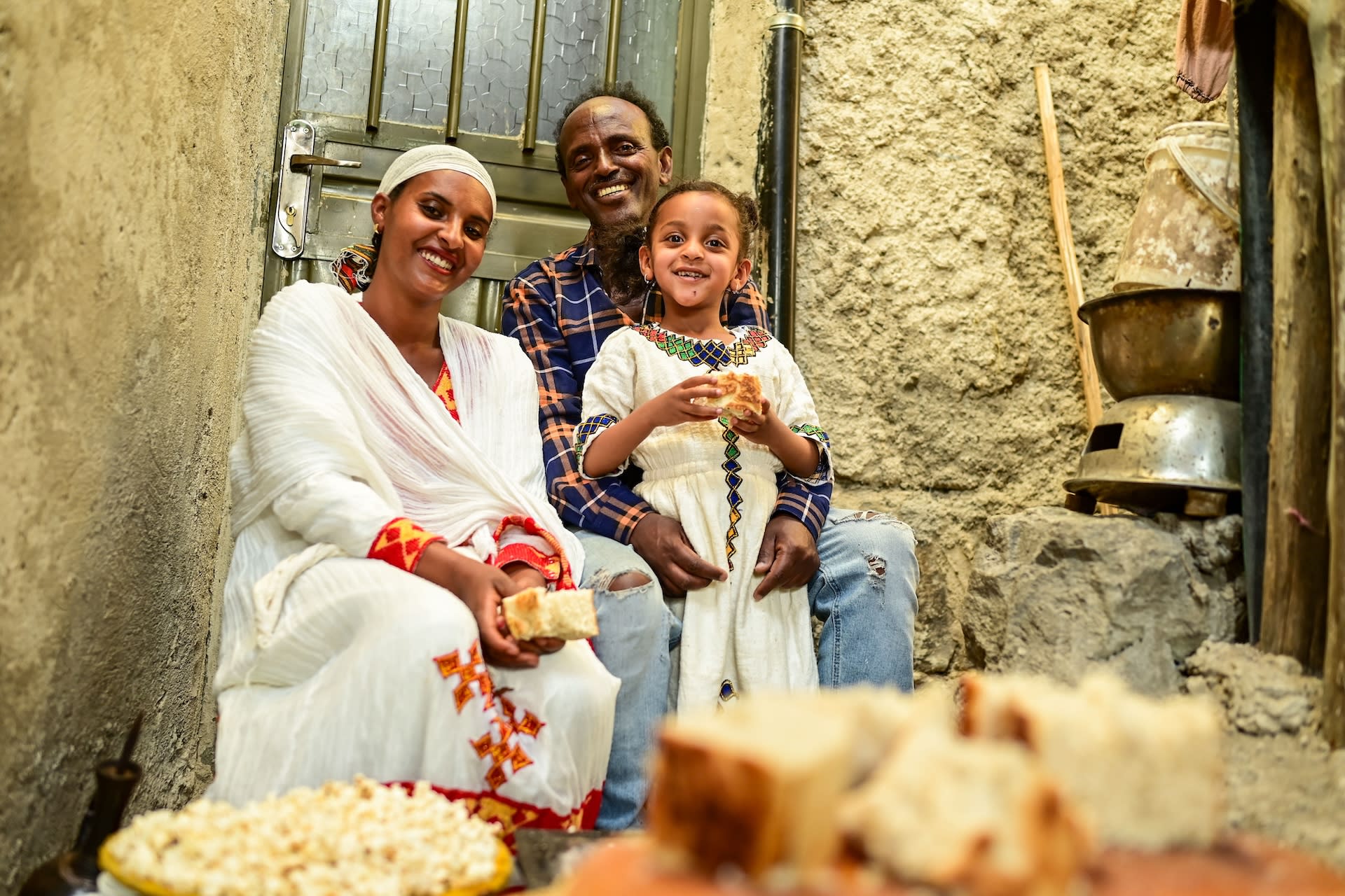 Anteneh, Tsehaynesh and Elbetel sit in front of the green door to their home. They are smiling and holding pieces of defo dabo, an Ethiopian holiday bread.