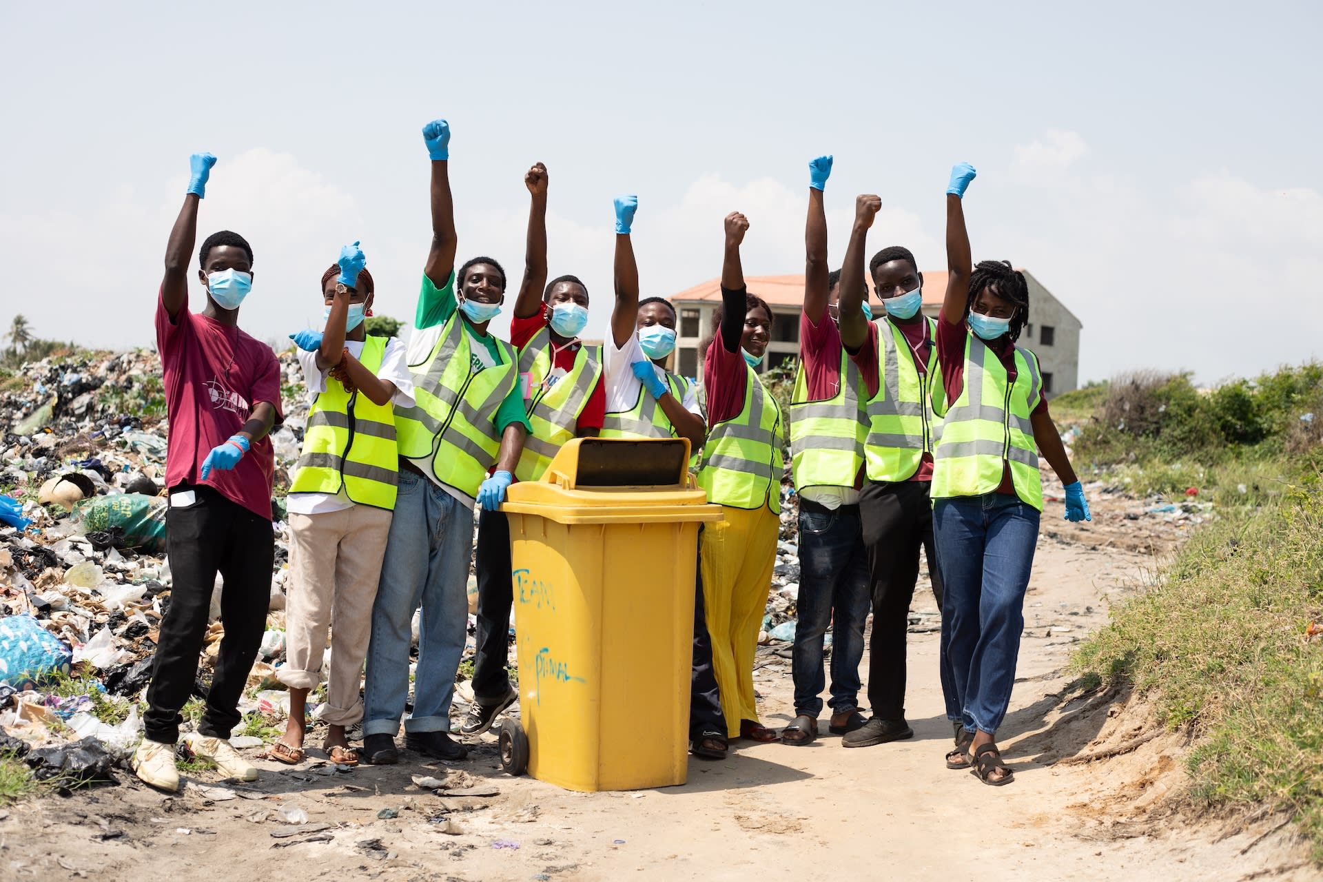 A group picture of Team Optimal, a recycling business started in Ningo, Ghana by a Compassion alumni. They are standing at a dumpsite, posing and smiling in excitement. A yellow recycle bin is in the middle of the group.