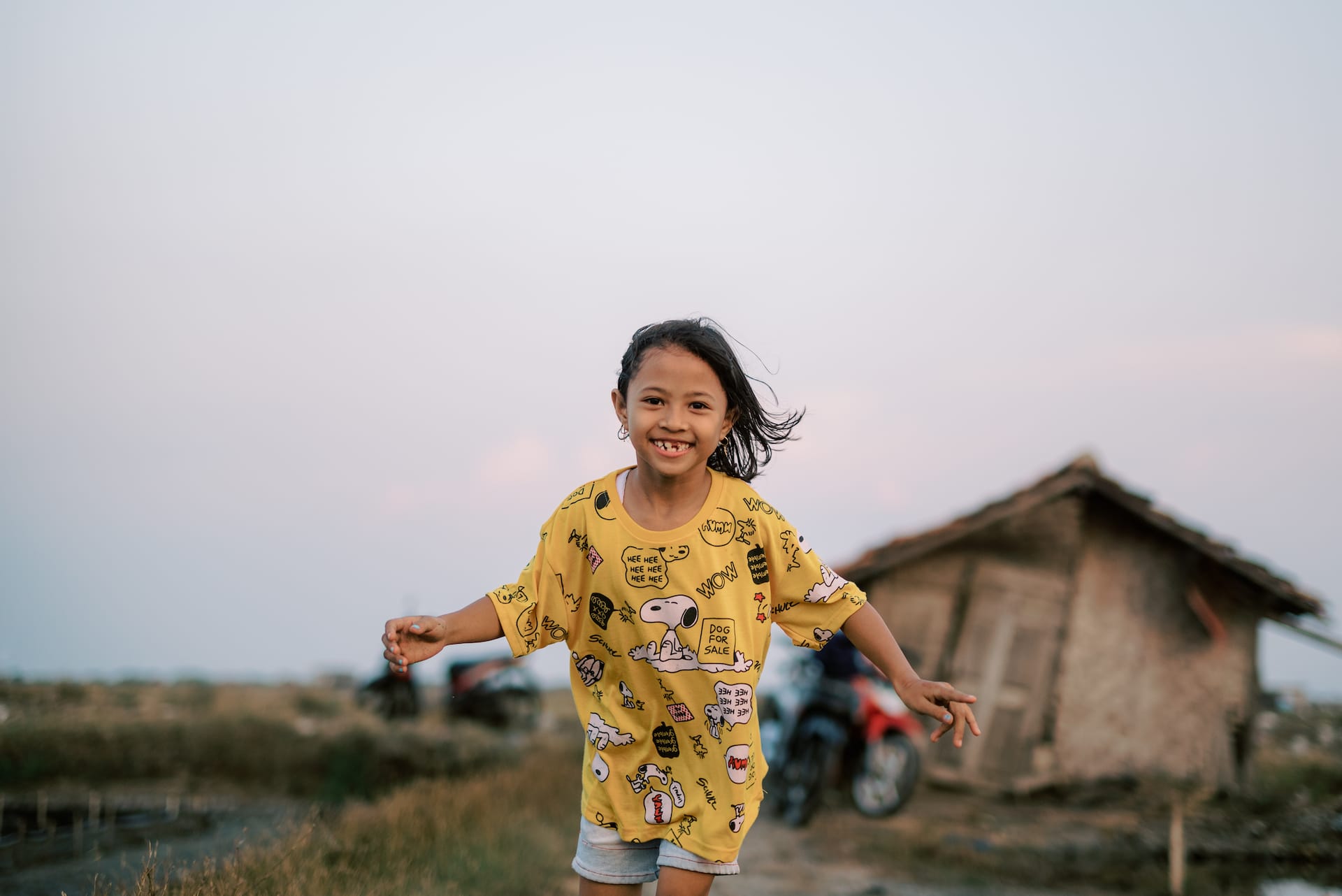 Filia, an 8-year-old girl in Indonesia, is wearing a yellow shirt and shorts. She is standing outside her home.