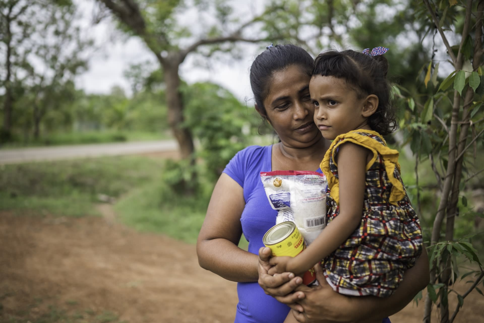 Ranuki is carried by her mother, who is holding some items from the food pack they received from the center. They standing by the side of the road next to where the local child development center is. Ranuki's mother is wearing a blue shirt and colorful skirt. Ranuki is wearing a printed dress.