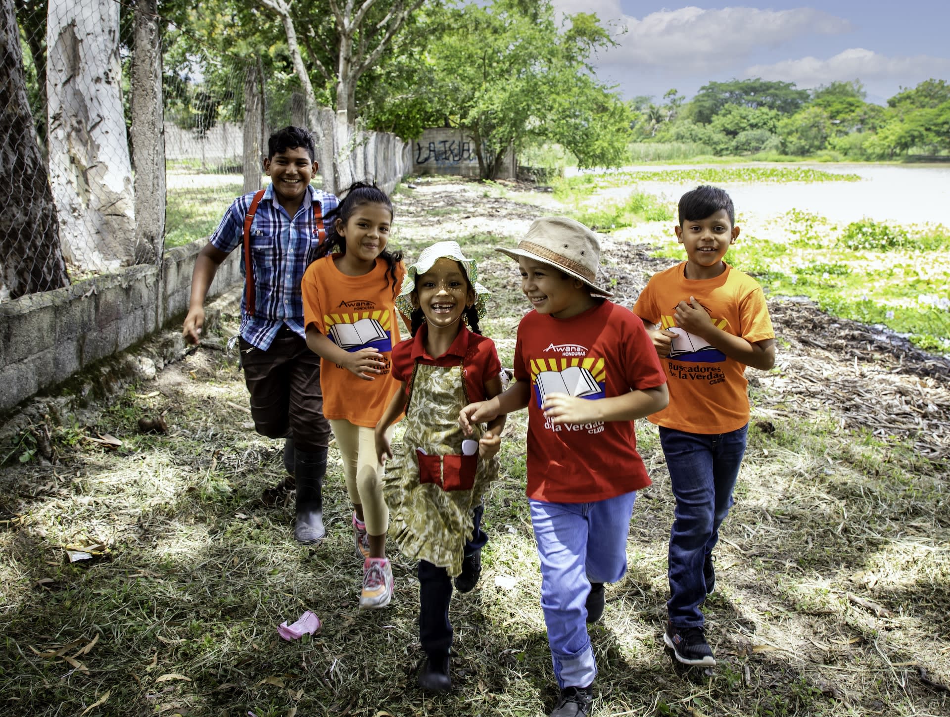 Five children running alongside a tall fence with smiles on their faces.
