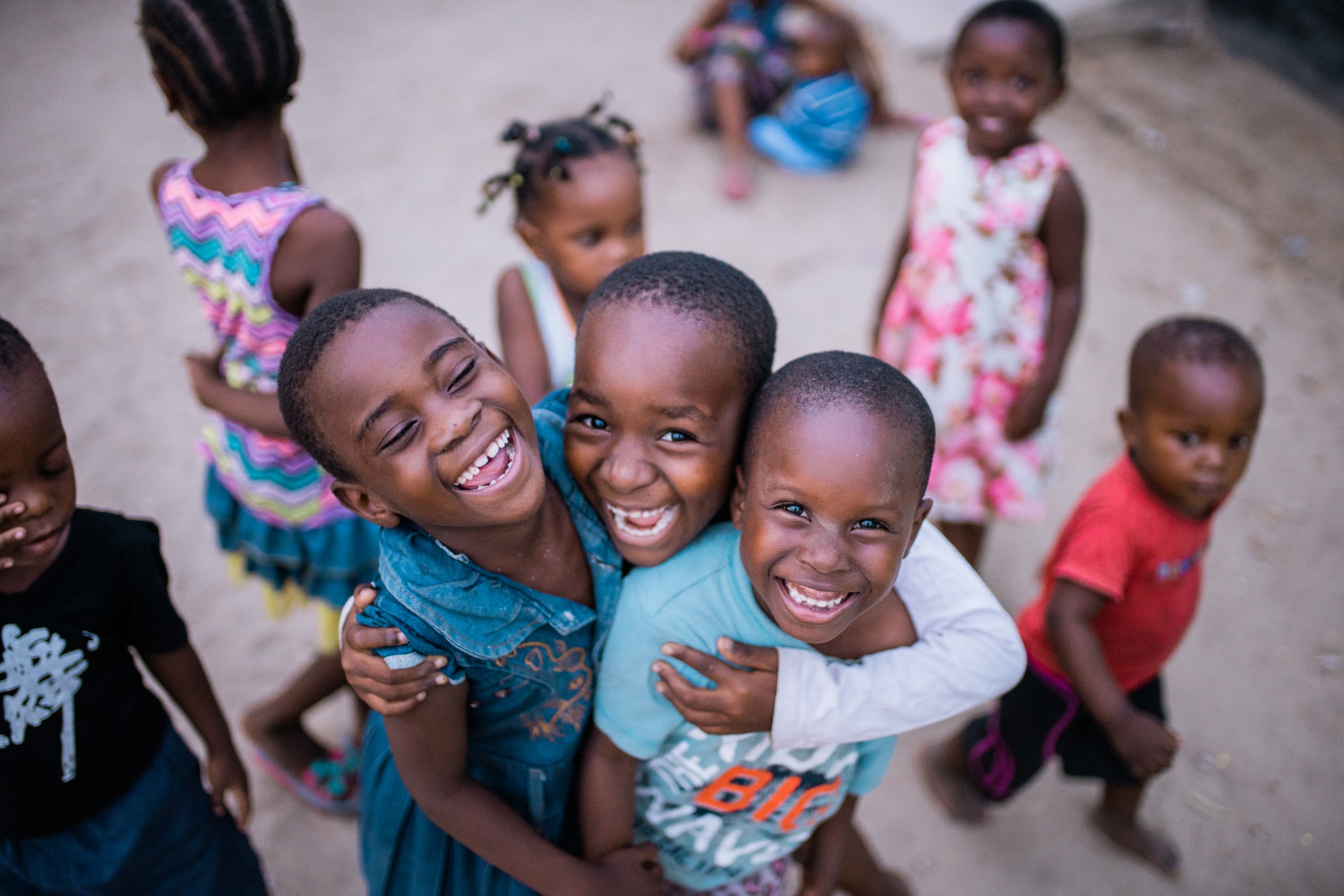 Three children laugh together in amidst a group of children playing outside.