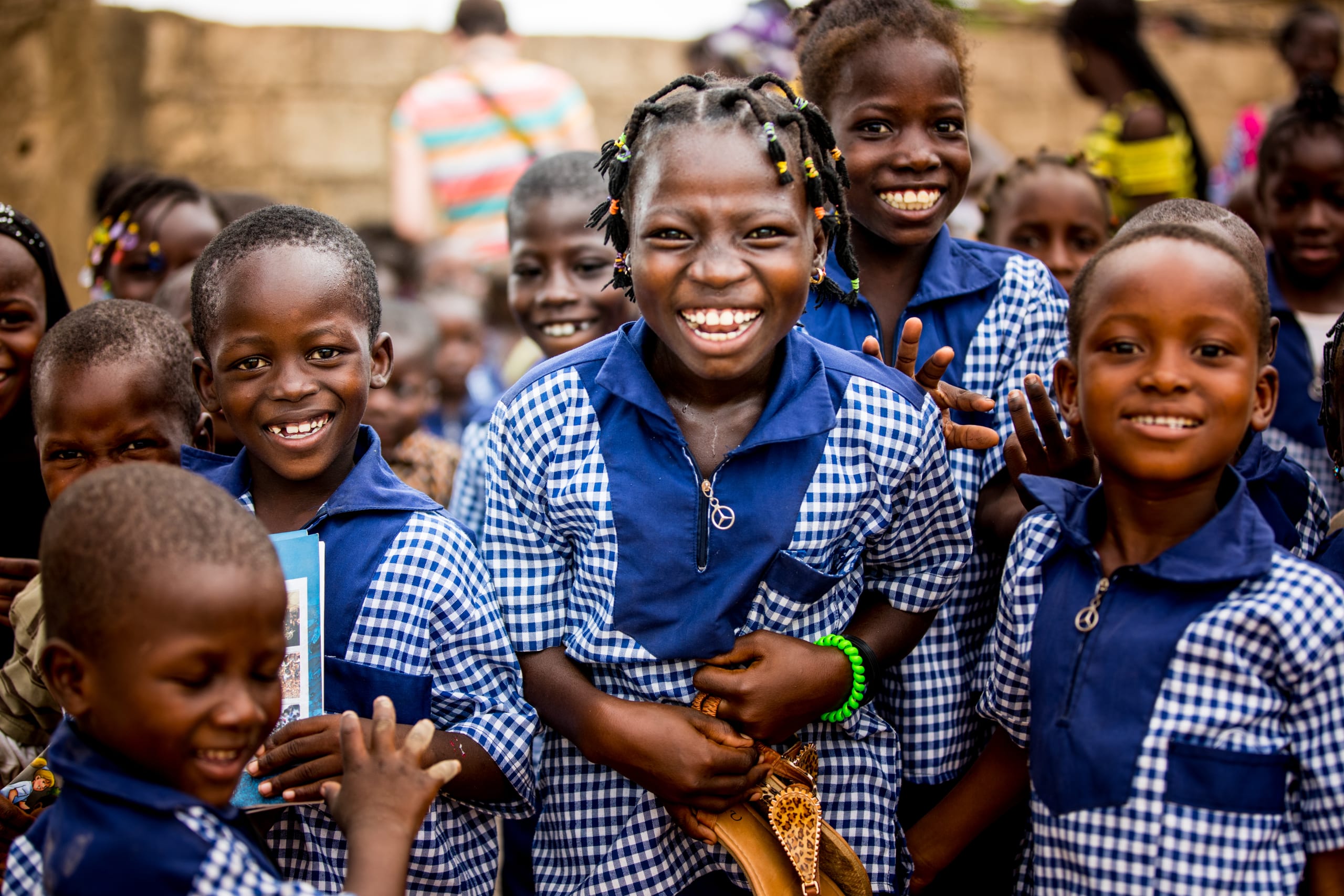 A group of children from Burkina Faso smile and laugh. They are wearing blue and white school uniforms and are standing in front of a Compassion centre building