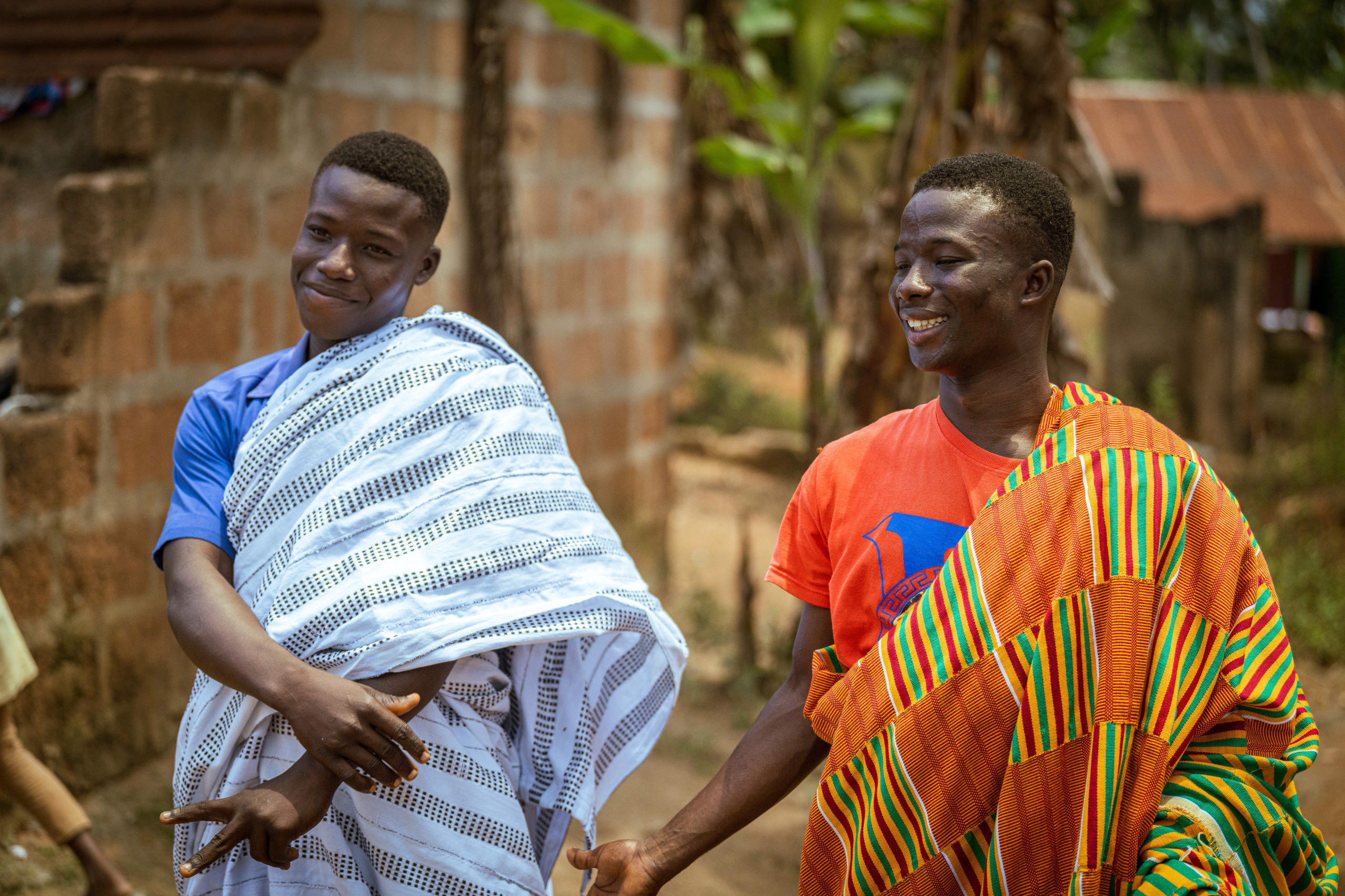 Two young boys stand and smile. They are wearing colourful kente cloth.