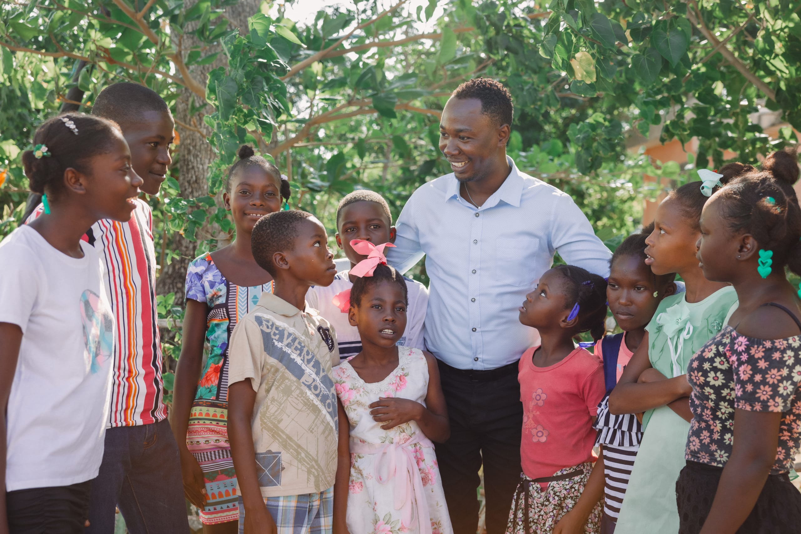 A man in a blue button-up shirt stands among children. They are smiling.